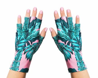 Puede incluir: Un par de guantes sin dedos con un vibrante estampado tropical de hojas de monstera y palmera en verde oscuro sobre un fondo rosa claro. Los guantes cubren las muñecas y dejan los dedos al descubierto para una total destreza manual.