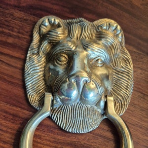 May include: A brass door knocker shaped like a lion's head with a ring handle. The lion's head is detailed with a mane, whiskers, and a textured surface.