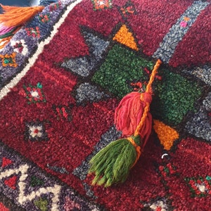 May include: A red, green, and orange patterned throw pillow with a fringe tassel. The pillow has a geometric design and is made of a soft, textured fabric.