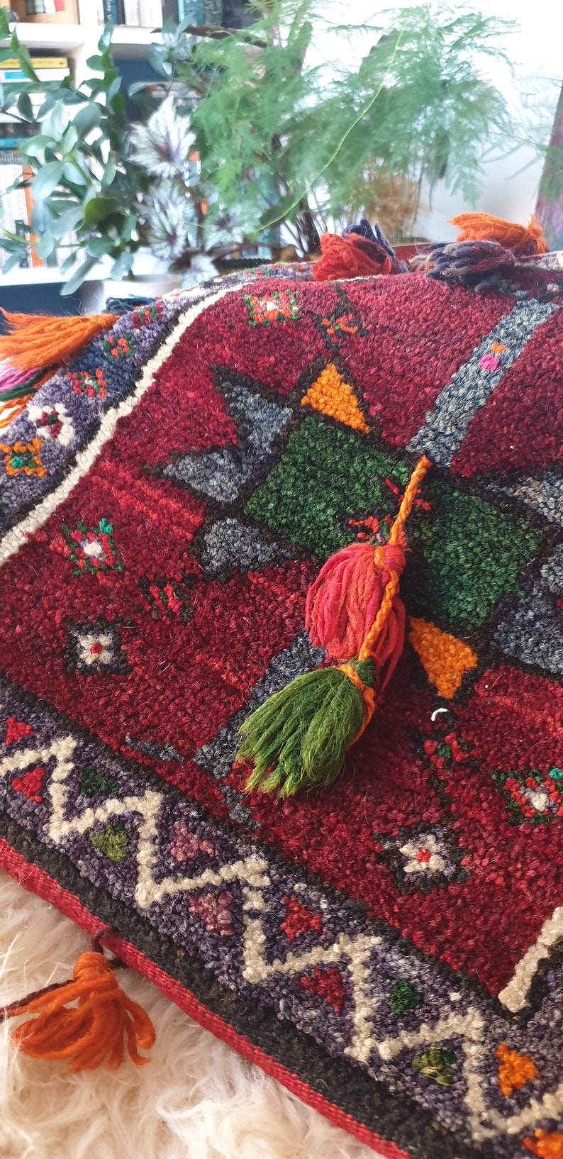 May include: A red, green, and orange patterned throw pillow with a fringe tassel. The pillow has a geometric design and is made of a soft, textured fabric.
