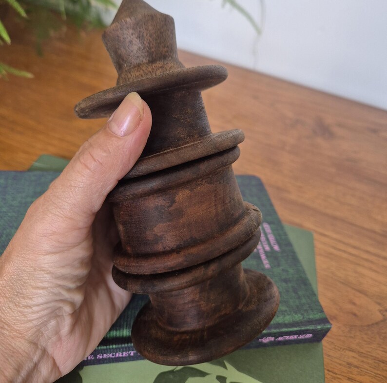 May include: A dark brown, hand-turned wooden object with a tiered, cylindrical structure featuring multiple horizontal ridges and a pointed top. The item has a smooth, polished finish with visible wood grain patterns and a wide, flat base.