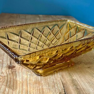 May include: Amber-coloured, rectangular glass serving bowl with a diamond pattern. The bowl has a square base and a decorative rim. The bowl is sitting on a wooden surface.