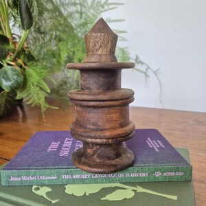May include: A dark brown wooden object with a stacked, cylindrical design featuring three distinct horizontal rings and a pointed conical top, resting on a purple book titled The Secret Language of Flowers by Jean-Michel Othoniel.