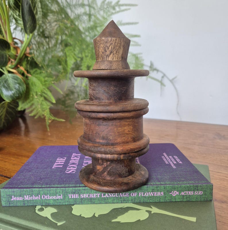May include: A dark brown wooden object with a stacked, cylindrical design featuring three distinct horizontal rings and a pointed conical top, resting on a purple book titled The Secret Language of Flowers by Jean-Michel Othoniel.