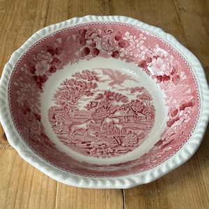 May include: A round, antique serving bowl with a red and white floral and pastoral design. The bowl features a scalloped edge and a detailed illustration of horses and a farmhouse. The rim has a white, rope-like texture.