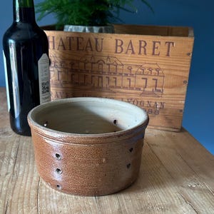 May include: A round, brown ceramic cheese mould featuring a series of small, evenly spaced drainage holes along its sides. The vessel has a smooth, light-coloured interior and a textured, darker exterior finish, designed for the process of cheese making.