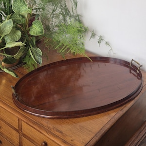 May include: An oval-shaped wooden serving tray with a polished dark brown finish and a smooth, reflective surface. The tray features two small, curved brass-toned metal handles attached to each end of the rim, providing a functional grip.