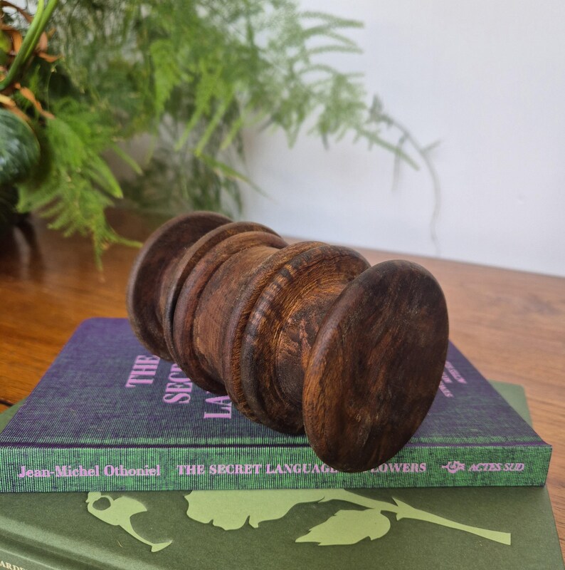 May include: A dark brown, cylindrical wooden spool with a ribbed, turned texture resting horizontally on a purple book titled The Secret Language of Flowers by Jean-Michel Othoniel, which is stacked atop a green book with a white floral graphic.