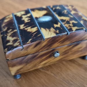 May include: A small, rectangular tortoiseshell box with a hinged lid and silver accents. The box has a decorative pattern and a silver knob on the front.