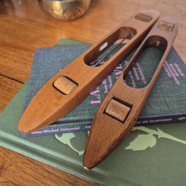 May include: Two elongated, boat-shaped wooden weaving shuttles with smooth, polished surfaces. Each shuttle features a central open cavity containing a small, rotating wooden bobbin. The wood grain is visible, showing natural brown tones and textures.