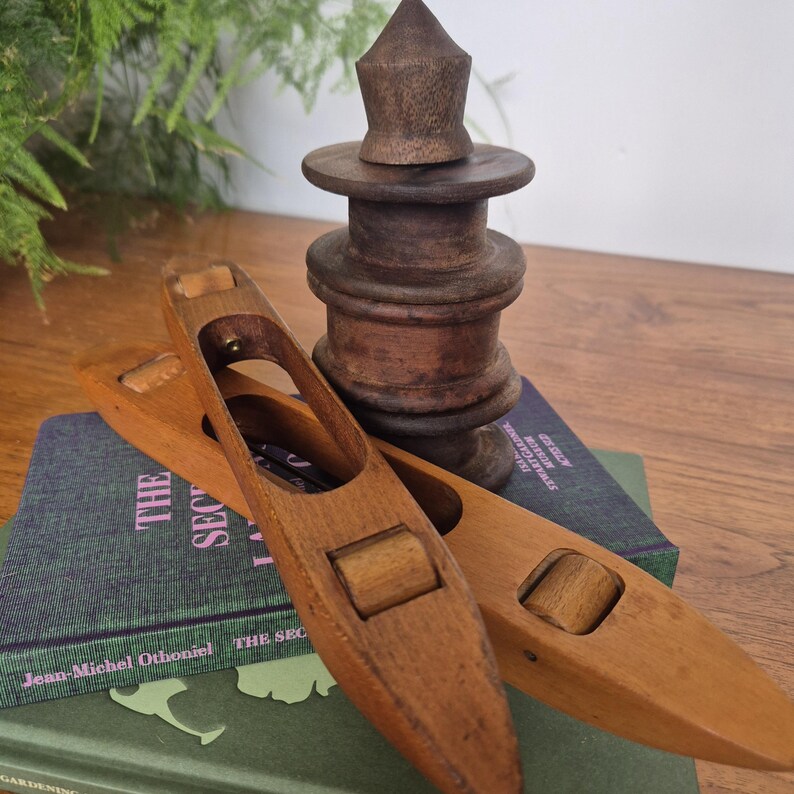 May include: Two wooden weaving shuttles with central rollers and a dark brown wooden bobbin stand. The shuttles are light brown with elongated openings. The bobbin stand features a tiered, cylindrical design with a pointed top finial.