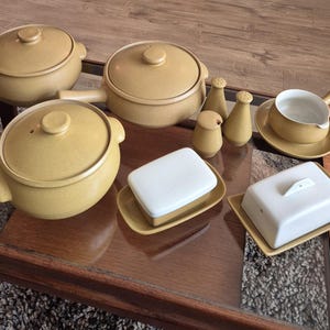 May include: A collection of tan ceramic cookware and serving dishes. Includes lidded pots, a saucepan, salt and pepper shakers, a gravy boat, and two butter dishes with white lids. The set is arranged on a glass-topped table.