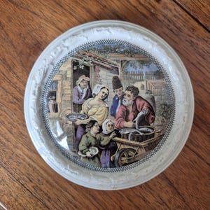 May include: A white decorative plate with a detailed, colourful illustration of a market scene. The plate features a raised, textured border and a detailed depiction of people, buildings, and a cart filled with fish.