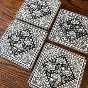 May include: Four square ceramic tiles featuring a black and white floral pattern. Each tile displays a central diamond shape filled with detailed flower illustrations, surrounded by a decorative border of repeating leaf and vine motifs.