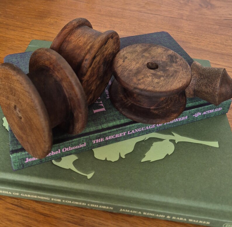 May include: Three dark brown wooden spools with a smooth, polished finish and a central hole, resting on top of two stacked hardcover books. The top book is green with the text The Secret Language of Flowers and the bottom book is dark green.