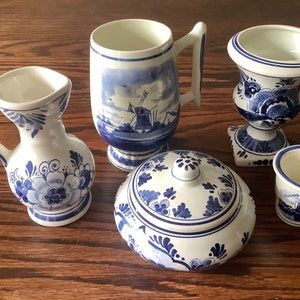 May include: A selection of white and blue Delftware pottery. The set comprises a small jug, a mug with a windmill scene, a lidded jar, a small vase, and a small cup with a windmill illustration.