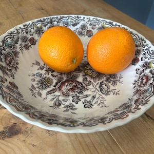May include: A white ceramic serving bowl with a floral pattern, holding two oranges. The oranges are bright orange and have a textured peel. The bowl has a scalloped edge and a detailed floral design.
