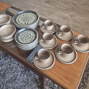 May include: A ceramic dinnerware set featuring stacks of plates with a dark brown rim and a central white teardrop pattern, alongside plain beige bowls and matching cups and saucers. The pieces have a matte, earthy finish and a consistent design.