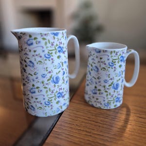 May include: Two white ceramic jugs with a blue and white floral pattern. The larger jug has a handle on the right side and the smaller jug has a handle on the left side.