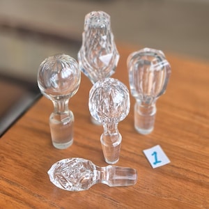 May include: Five clear glass decanter stoppers with various shapes and faceted or ribbed textures. Four stoppers are standing upright, and one is lying horizontally. A small white paper square with the handwritten number 1 is visible.