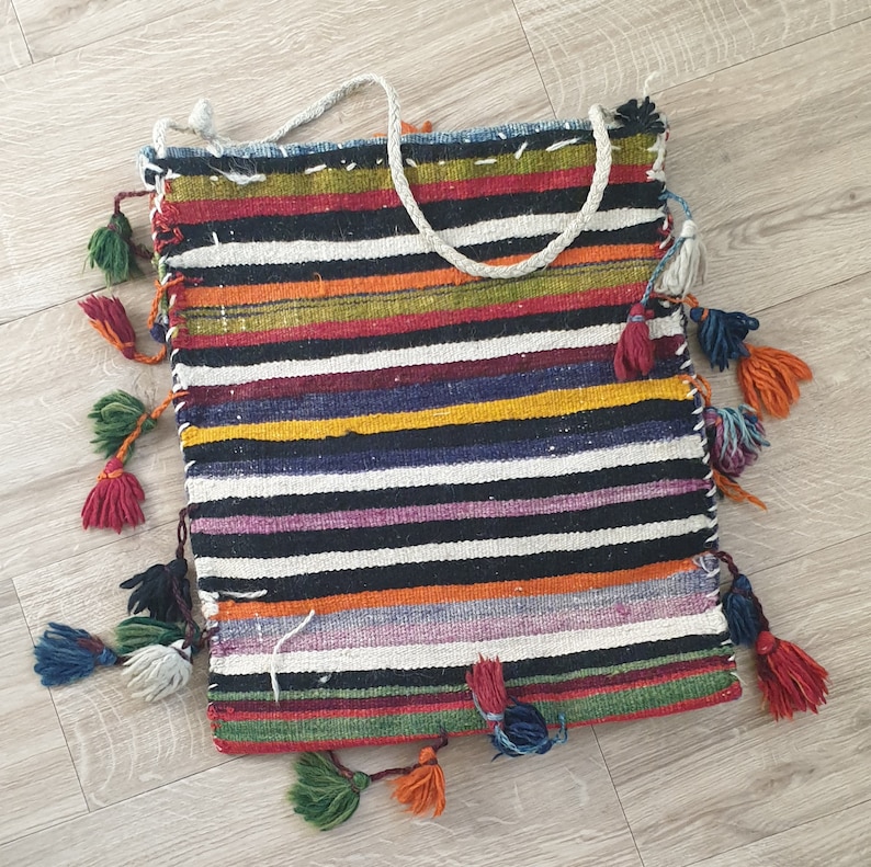 May include: A colorful striped woven bag with fringe tassels. The bag is made of a thick, textured fabric and has a black and white striped pattern with accents of red, yellow, green, and purple.
