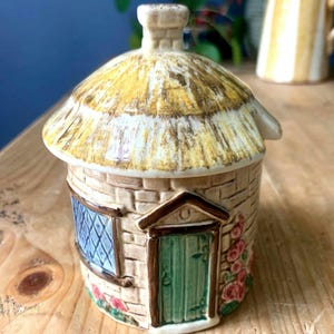 May include: A ceramic cottage-shaped container with a thatched roof and a chimney. The container has a green door, a blue window, and is decorated with pink roses. The walls are made of beige bricks.