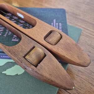 May include: Two wooden weaving shuttles with smooth, polished surfaces and integrated rollers. Each shuttle features an elongated, tapered shape with a central open slot and a small, cylindrical wooden wheel set into the body for smooth movement.