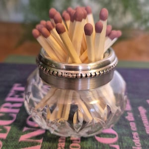 May include: A glass match holder with a silver band, containing wooden matches. The match heads are reddish-brown. The holder is on a patterned surface. The glass has a cut-glass design.