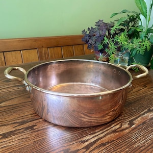 May include: A copper cooking pot with brass handles. The pot is round with a wide, shallow design. The copper has a warm, reddish-brown hue, and the brass handles curve outward. The pot sits on a wooden surface.