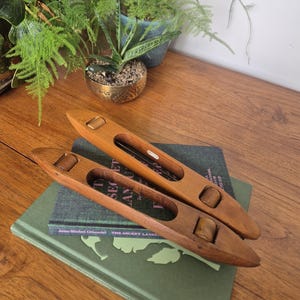 May include: Two smooth, light brown wooden weaving shuttles with tapered ends and central cutouts. Each shuttle features two small, cylindrical wooden rollers positioned within the frame. The shuttles are resting on a dark green hardcover book.