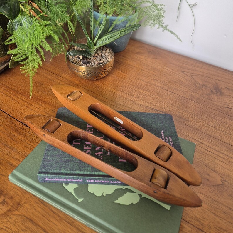 May include: Two smooth, light brown wooden weaving shuttles with tapered ends and central cutouts. Each shuttle features two small, cylindrical wooden rollers positioned within the frame. The shuttles are resting on a dark green hardcover book.
