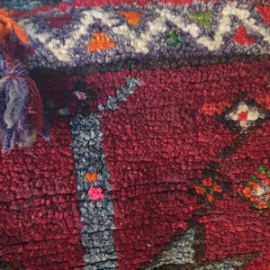May include: A close-up of a colorful woven rug with tassels. The rug has a red, green, blue, and orange pattern. The tassels are red and orange.