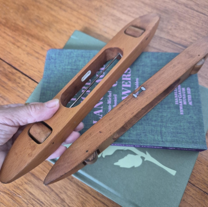 May include: Two elongated, smooth wooden weaving shuttles with tapered ends. One shuttle features a central open slot containing a metal rod and a small white insert, while both have recessed wooden rollers at each end for thread management.