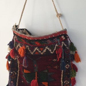 May include: A woven tapestry bag with a red, blue, green, and orange pattern. The bag has a long rope strap and is decorated with tassels.