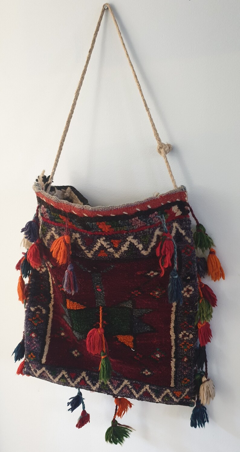 May include: A woven tapestry bag with a red, blue, green, and orange pattern. The bag has a long rope strap and is decorated with tassels.