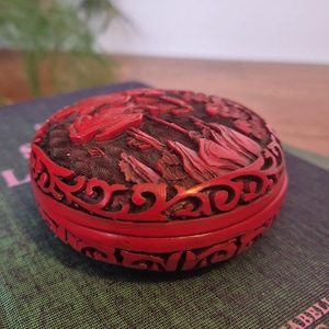 May include: A round, red lacquered box with detailed carvings. The lid displays a relief scene with figures and foliage. The box has a dark brown base and a decorative, openwork border. The design is ornate and detailed, showcasing fine craftsmanship.
