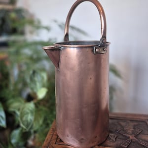 May include: A copper jug with a spout and a curved handle. The jug is cylindrical and made of copper with a polished finish. The handle is attached to the top with rivets. The jug is displayed on a wooden surface.