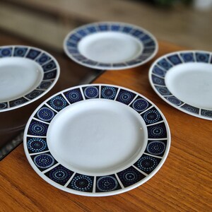 May include: Four white ceramic plates with a blue and white geometric pattern. The plates are arranged in a circle on a wooden surface.