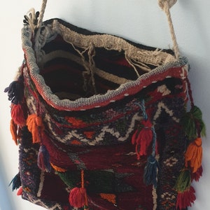May include: A woven tapestry bag with a long, knotted rope strap. The bag is decorated with colorful tassels and intricate patterns in shades of red, blue, green, and orange.