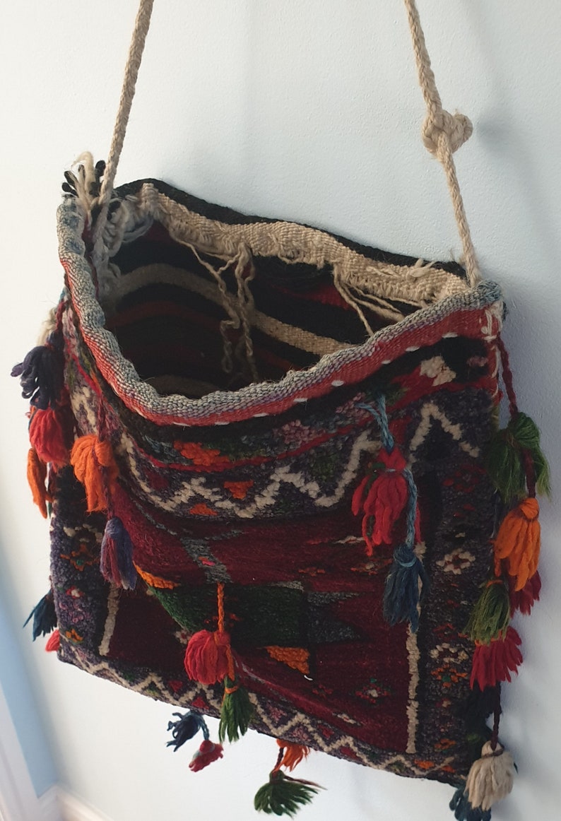 May include: A woven tapestry bag with a long, knotted rope strap. The bag is decorated with colorful tassels and intricate patterns in shades of red, blue, green, and orange.