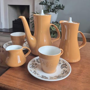 May include: A retro orange ceramic tea set, including a teapot, milk jug, sugar bowl, and cups with saucers. The cups and saucers have a white interior and a floral pattern. The set is on a wooden table.
