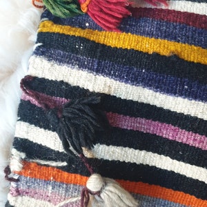 May include: A colorful striped woven bag with multiple tassels in various colors, including green, red, white, and black. The bag is made of a thick, textured fabric and has a fringed edge.