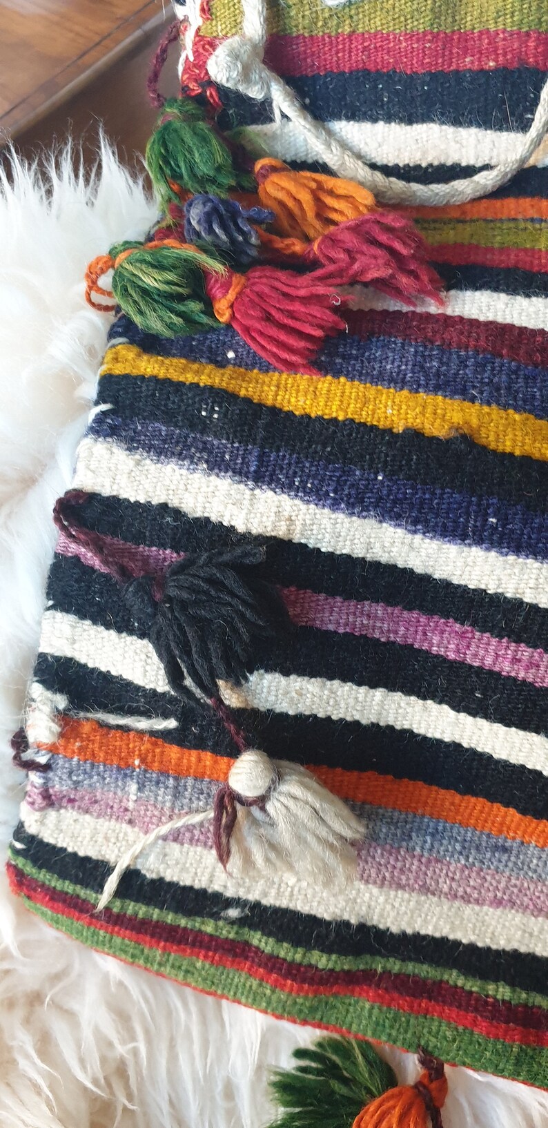 May include: A colorful striped woven bag with multiple tassels in various colors, including green, red, white, and black. The bag is made of a thick, textured fabric and has a fringed edge.