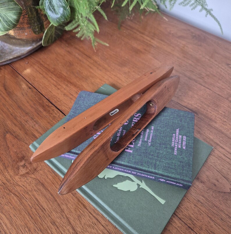 May include: Two smooth, polished wooden weaving shuttles with pointed ends and central openings. The shuttles feature a warm, medium-brown wood grain and are positioned at an angle, resting on top of a green hardcover book with visible text.