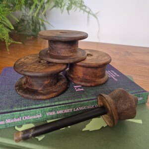 May include: Three dark brown wooden spools with a smooth finish and a central hole, arranged in a cluster. A single wooden tool with a long black handle and a flared brown top piece lies horizontally in front of the spools on a book.