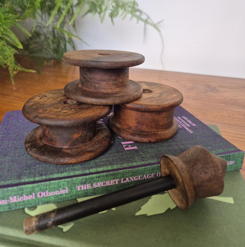 May include: Three dark brown wooden spools with a smooth finish and a central hole, arranged in a cluster. A single wooden tool with a long black handle and a flared brown top piece lies horizontally in front of the spools on a book.