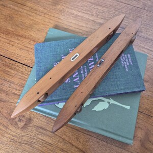 May include: Two elongated, pointed wooden weaving shuttles with smooth, light brown surfaces. Each shuttle features a central opening, small metal components, and a small wheel visible at one end, designed for use in manual loom weaving processes.