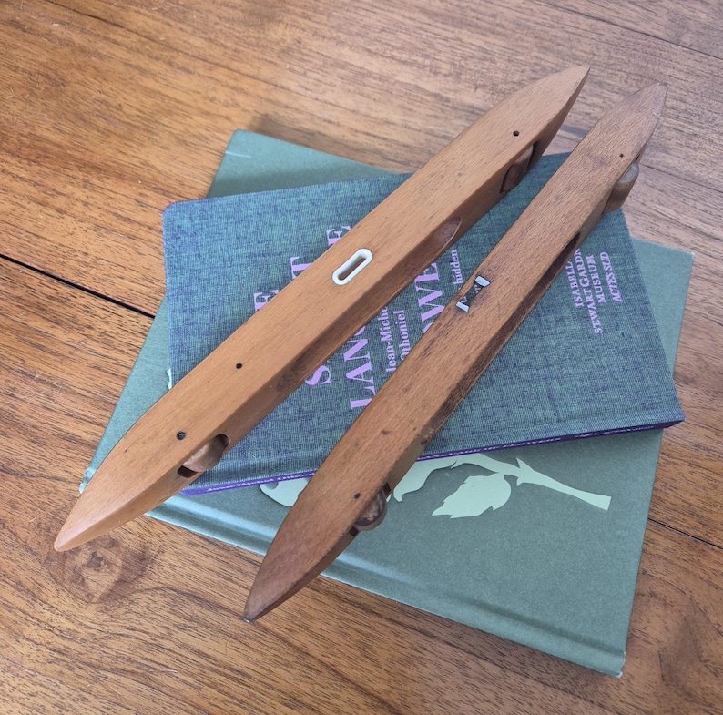 May include: Two elongated, pointed wooden weaving shuttles with smooth, light brown surfaces. Each shuttle features a central opening, small metal components, and a small wheel visible at one end, designed for use in manual loom weaving processes.