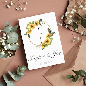 Puede incluir: Una invitación de boda blanca con una corona de girasoles y detalles dorados. Las iniciales "T" y "J" están en el centro de la corona. El texto "Taylor & Joe" y la fecha "13.08.2023" están debajo de la corona.