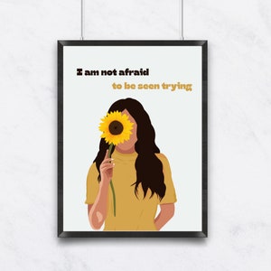 May include: A woman with long brown hair holds a sunflower in front of her face. The sunflower is yellow with a dark brown center. The background is white with the text "I am not afraid to be seen trying".